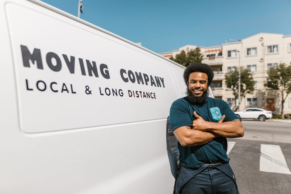 A smiling male mover with dark curly hair and a full beard, wearing a dark blue t-shirt, stands outdoors next to a white moving van. The van displays the text 'MOVING COMPANY LOCAL & LONG DISTANCE' in