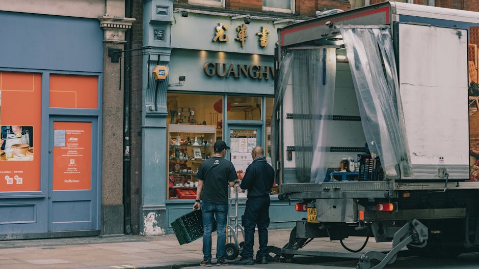 Two men stand outside the storefront of a shop named Guangh, engaged in a discussion during a home relocation or furniture transport process. One man is holding a large black crate, while the other is