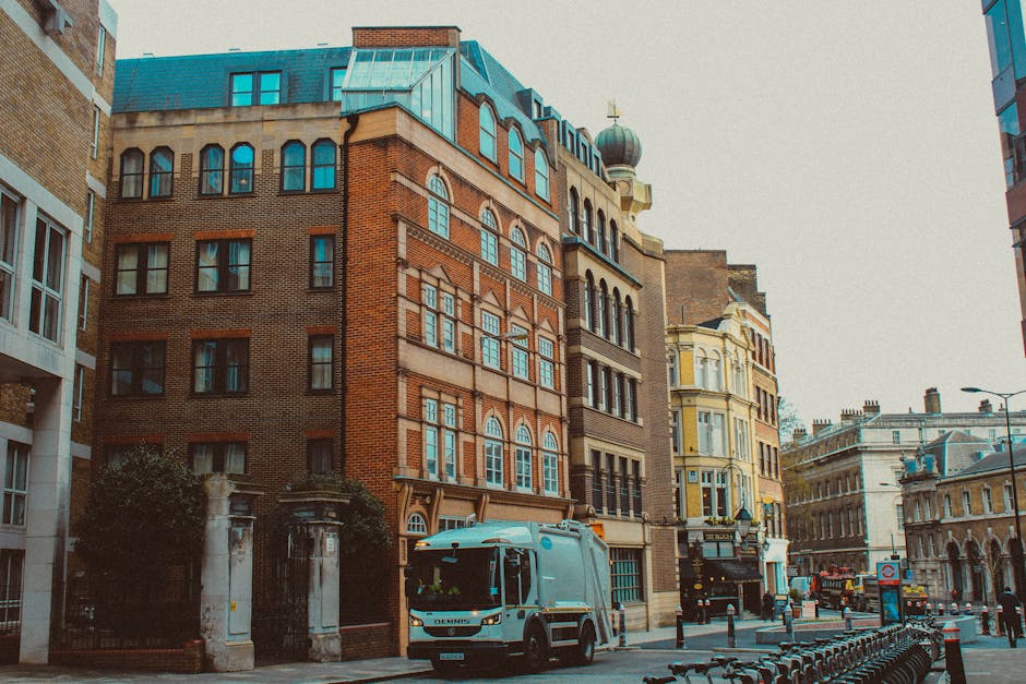 The image depicts a city street scene during daytime with a street-facing building made of red and beige bricks, featuring multiple large windows with decorative arches, and a modern blue glass roofto